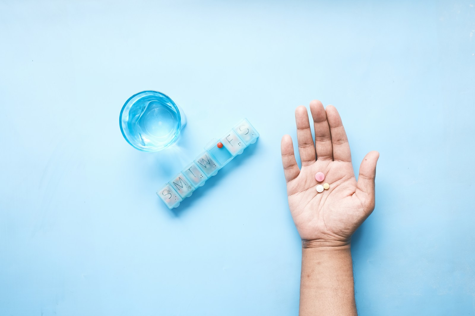 Hand holding medication pills with a glass of water and a weekly pill organizer